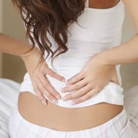 Woman sitting down on edge of a bed holding her lower back with both hands