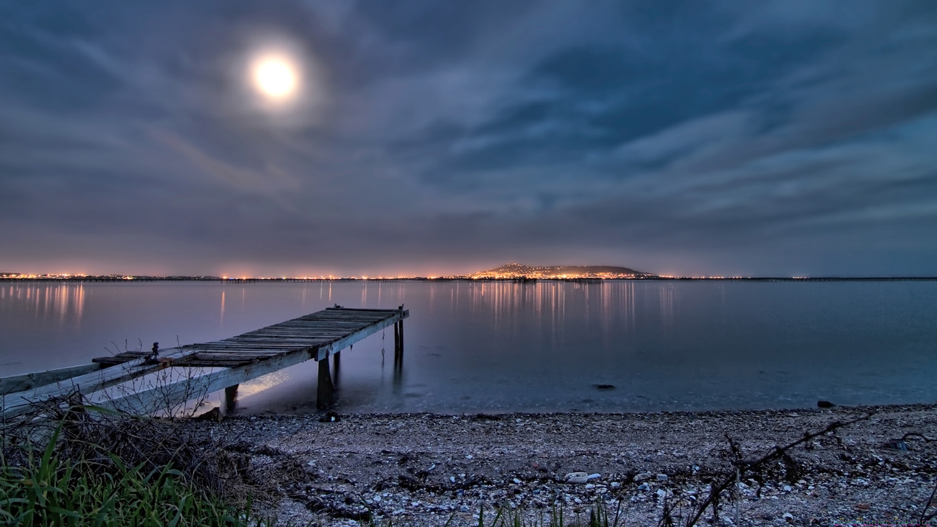 Pier protruding into a lake across from civilization during early evening with moon visible